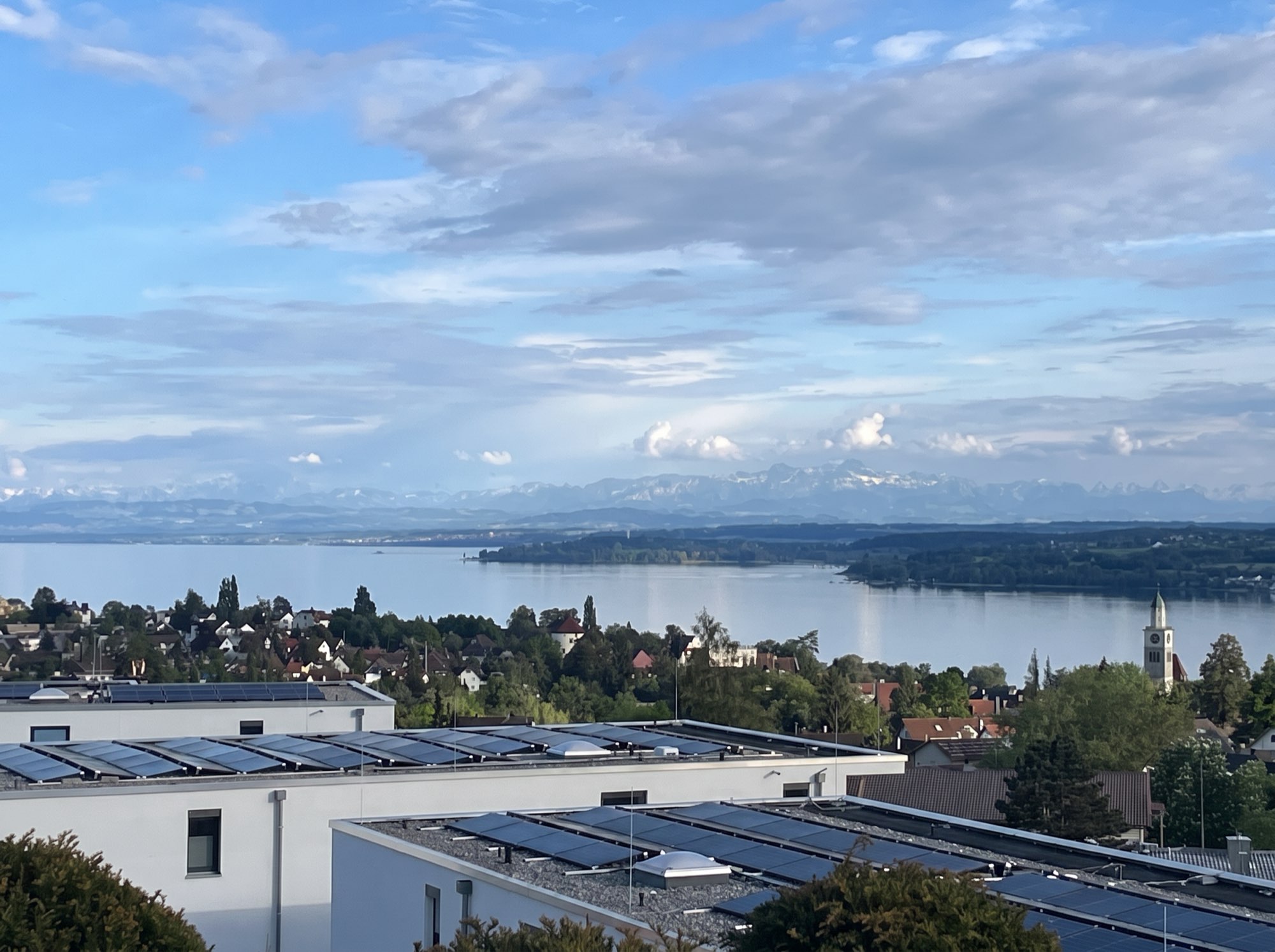 Blick von der Dachterrasse des Q5 über die Dächer Überlingens auf den Bodensee, dahinter die Alpenkette im Abendlicht.